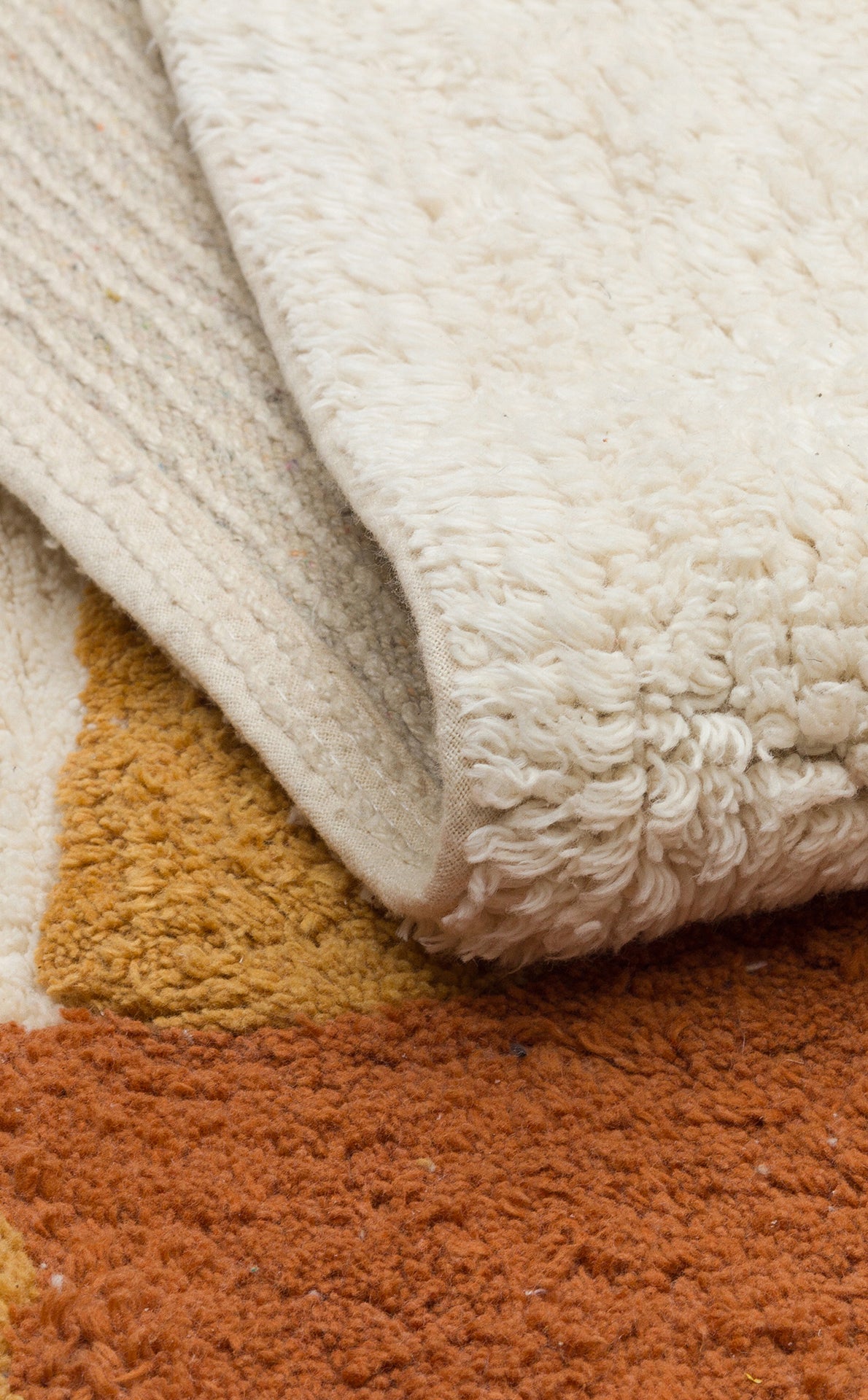 A close-up of the Rainbow Nap - Washable Shaggy - CBN08 rug, cream-colored and folded over a patterned rug with mustard yellow and rust orange accents. Both have thick, tufted surfaces, perfect for cozy kids' playroom setups.
