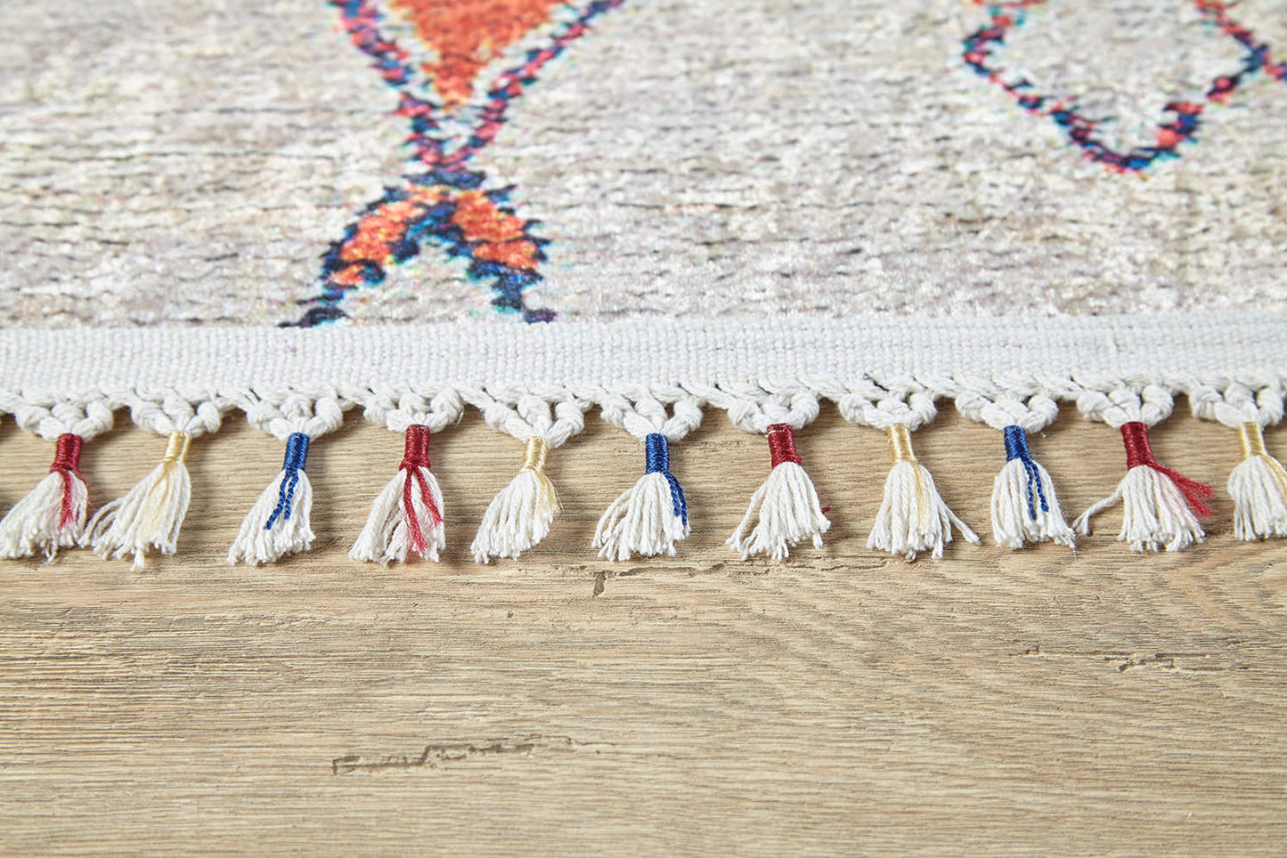 Close-up of the Rainbow Rhapsody - Washable Rug - JR5060 (Custom Size) edge, featuring knotted white fringe with some tassels in blue or red, on a light wood floor. Its colorful geometric pattern is ideal for high-traffic areas.