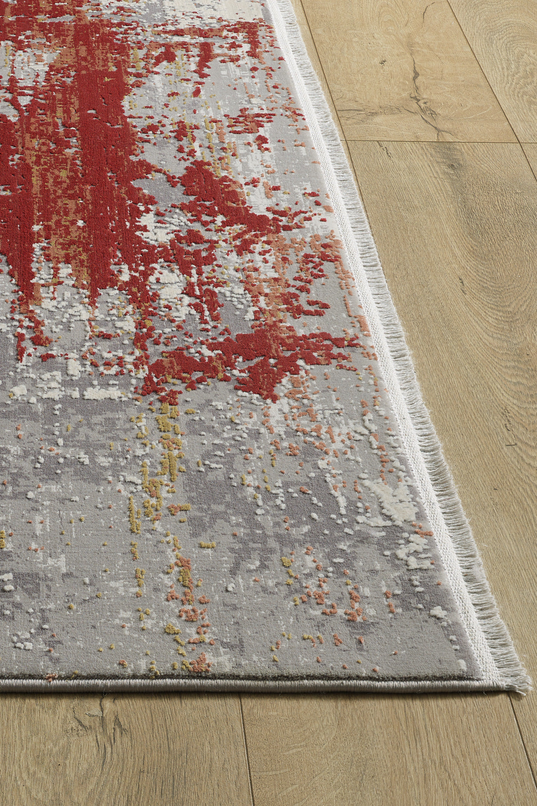 A close-up of the Red Strokes Premium Rug - 1052EB on a wooden floor, displaying an abstract rectangular design in red, gray, white, and yellow with fringe along the shorter edges.