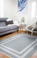 A modern living room features a gray sofa, patterned armchair, and the Refined Edge - Washable Rug - JR573 (Custom Size), ideal for high-traffic areas. A small round table with a face vase sits beneath blue leaf artwork on the white wall.