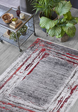 A modern living room featuring the Refined and Timeless - Washable Rug - JR1500 (Custom Size) in gray and red abstract design, a small metal table with decor, and two large potted plants on light wood flooring—ideal for busy spaces.