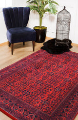 The Regal Accents - Washable Rug - JR36 (Custom Size) in a red pattern covers a wooden floor near a navy velvet chair, green potted plant, black fur rug, and an empty birdcage—ideal for high-traffic areas.