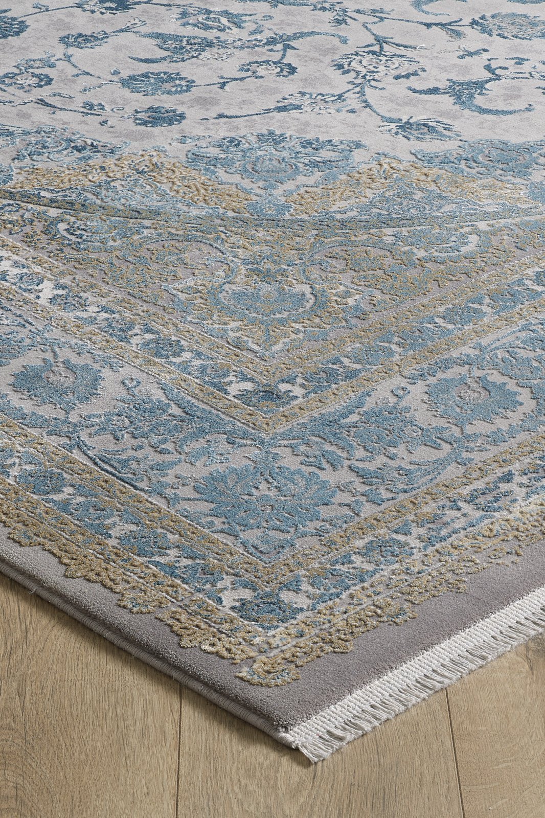 The Reimagined Heritage Modernised Medallion Rug - Sky - M642A showcases an intricate blue and gold pattern on a light gray background with white fringe, seen here in the corner placed on a light wood floor.