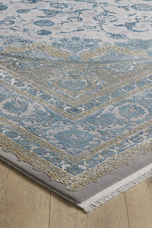 Load image 1 in gallery view. The Reimagined Heritage Modernised Medallion Rug - Sky - M642A showcases an intricate blue and gold pattern on a light gray background with white fringe, seen here in the corner placed on a light wood floor.