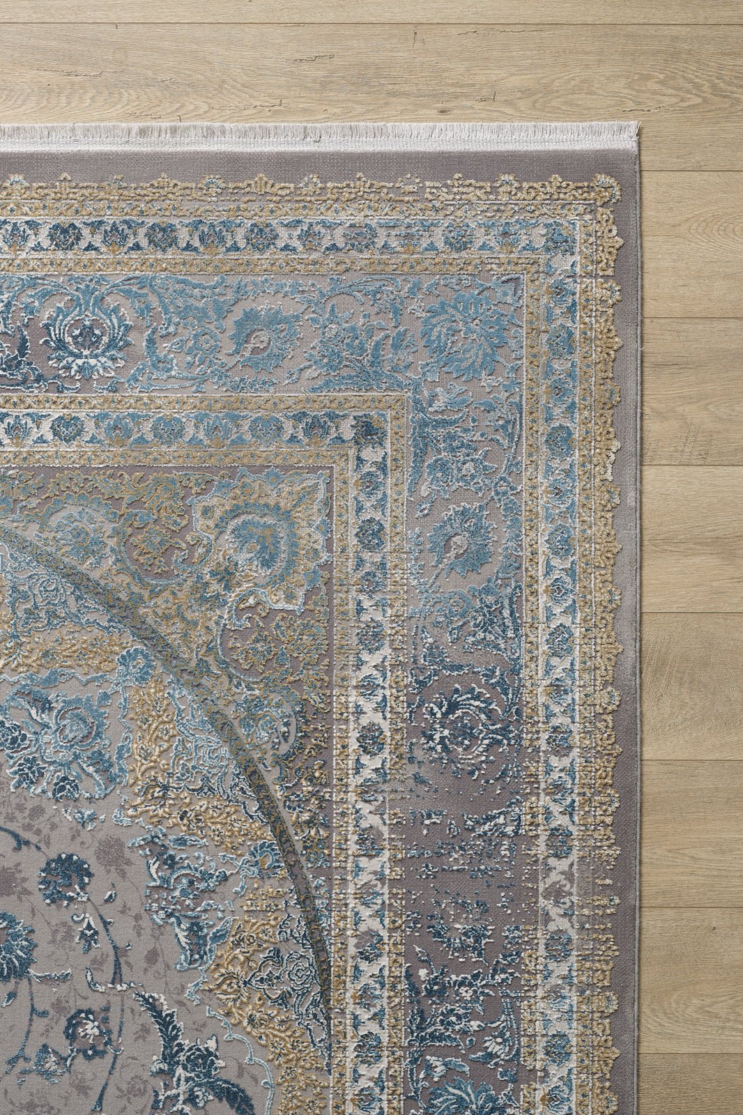 A close-up of the Reimagined Heritage Modernised Medallion Rug - Sky - M642A, featuring ornate blue, gold, and beige patterns with intricate borders and white fringe, displayed on a light wooden floor.