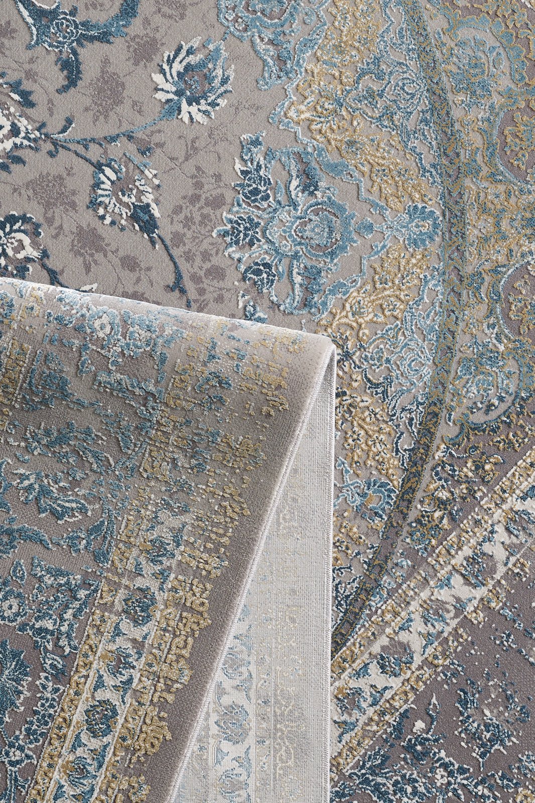 A close-up of the Reimagined Heritage Modernised Medallion Rug - Sky - M642A shows its ornate blue, gold, and gray floral and geometric patterns. One corner is folded over to display both sides of this elegant rug.