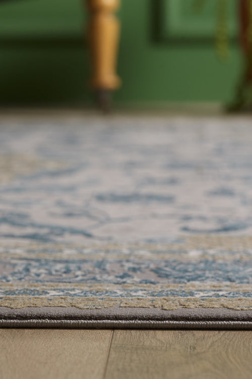 Load image 1 in gallery view. A close-up of the Reimagined Heritage Modernised Medallion Rug - Sky - M642A with blue and beige designs on a wooden floor, featuring a blurred wooden furniture leg and green wall in the background.