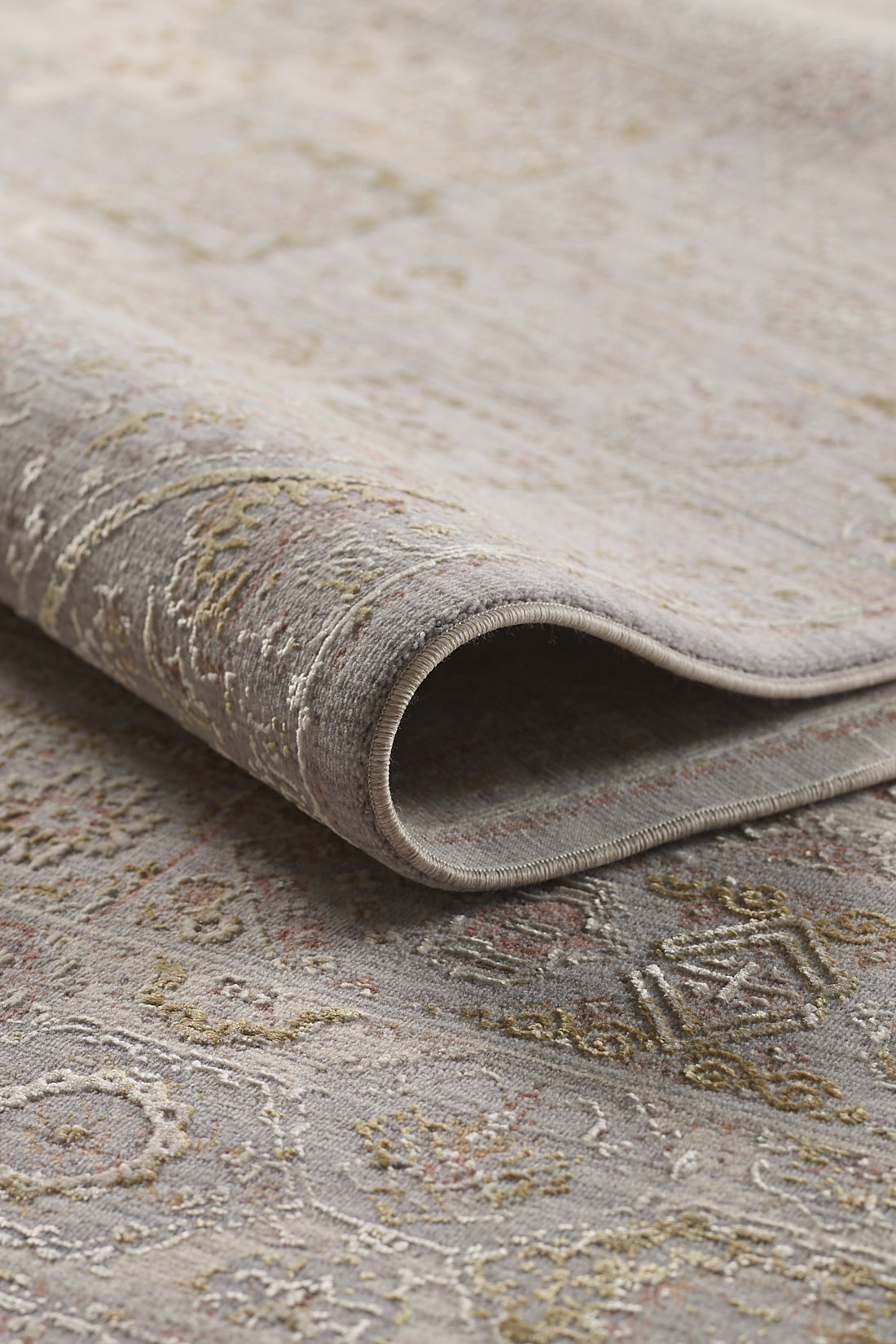 A close-up of the Revived Legacy Premium Rug - M616M, folded to showcase its intricate machine-made patterns in muted gray, beige, and gold tones with a detailed texture and soft woven surface.