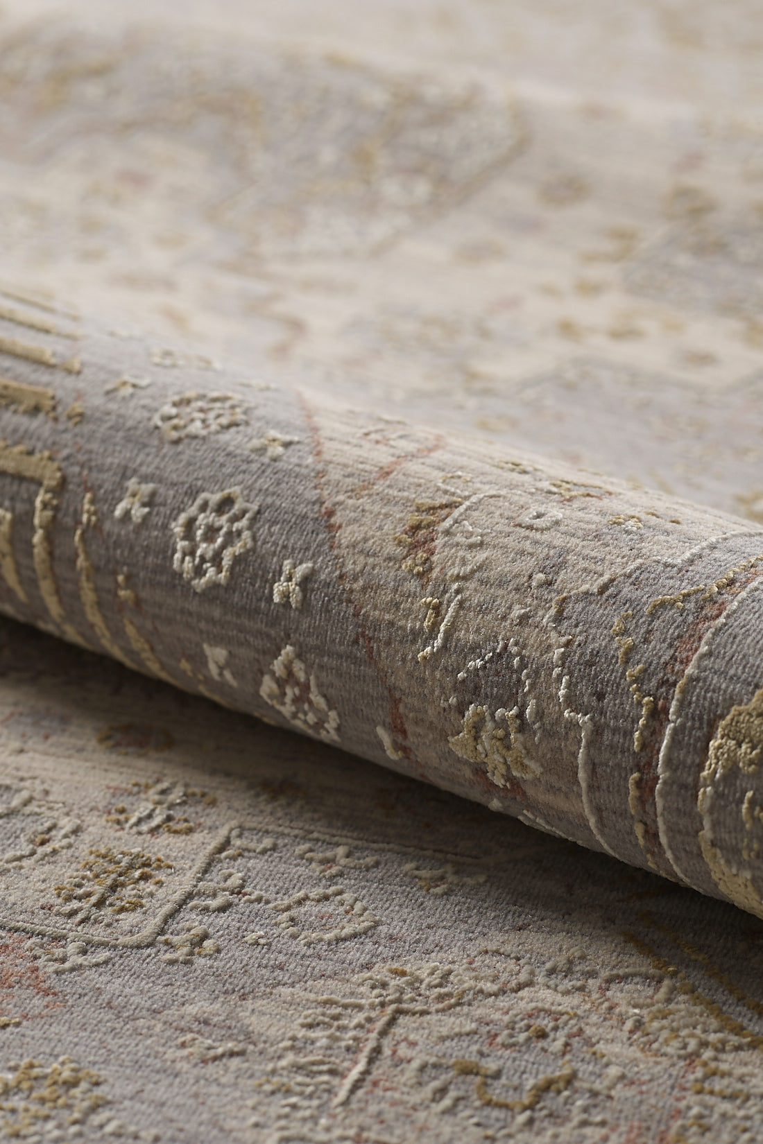 A close-up of the Revived Legacy Premium Rug - M616M in muted grey, beige, and gold tones highlights its textured fabric and intricate geometric and floral designs, with a portion of the rug rolled up.