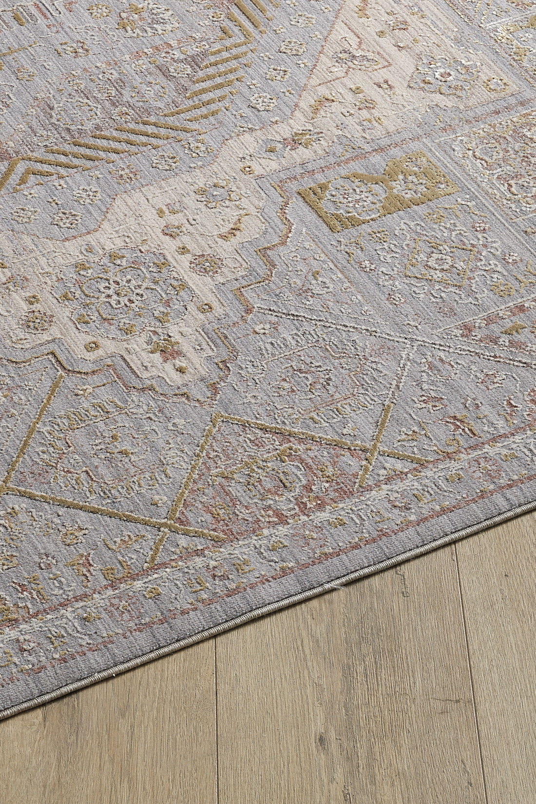 A close-up of the Revived Legacy Premium Rug - M616M, a viscose polyester piece with muted geometric patterns in light gray, beige, and gold tones, featuring floral accents and displayed on a light wood floor.