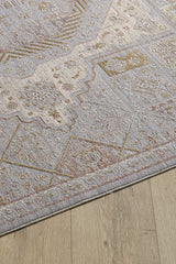 A close-up of the Revived Legacy Premium Rug - M616M, a viscose polyester piece with muted geometric patterns in light gray, beige, and gold tones, featuring floral accents and displayed on a light wood floor.