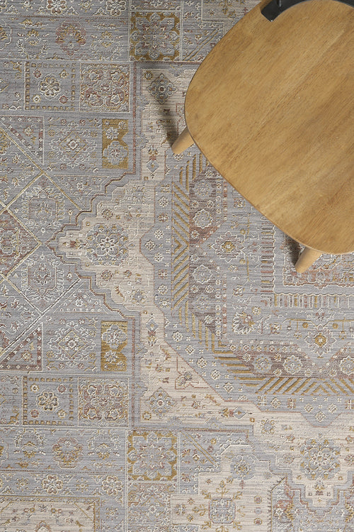 Load image 1 in gallery view. A portion of a wooden table rests on the Revived Legacy Premium Rug - M616M, displaying its intricate beige, gray, gold, and muted red floral patterns. The detailed machine-made design is highlighted in the corner of the image.