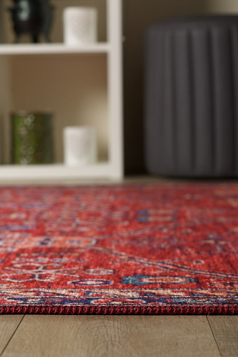 A close-up of the Rhapsody Authentic Red Washable Rug - LCC3033 (Outlet) US with a stylish pattern on a wooden floor; in the background, a white shelving unit holds candles and a green glass jar beside a dark gray ottoman—great for high-traffic areas.