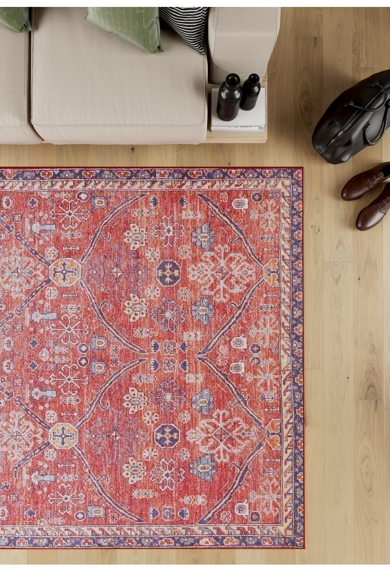 The Rhapsody Authentic Red Washable Rug (LCC3033) features blue and white accents and rests on a light wood floor near a beige sofa with green pillows, making it ideal for high-traffic areas. A side table holds two bottles, a black bag, and brown shoes.