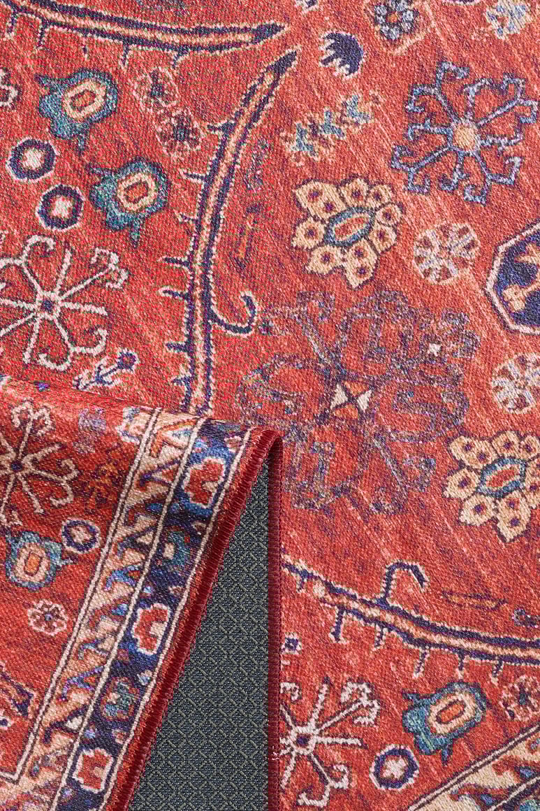 The Rhapsody Authentic Red Washable Rug - LCC3033 features intricate blue, cream, and gold floral and geometric designs. Its dark, diamond-patterned non-slip backing makes it ideal for high-traffic areas. One corner is shown folded over.