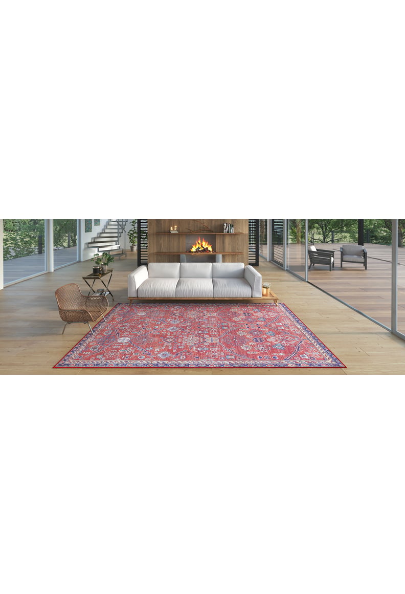 A modern living room features a white sofa, fireplace, wood coffee table, woven chair, and a Rhapsody Authentic Red Washable Rug - LCC3033 (Custom Size), perfect for high-traffic areas on light wood floors. Trees and patio furniture are visible outside.