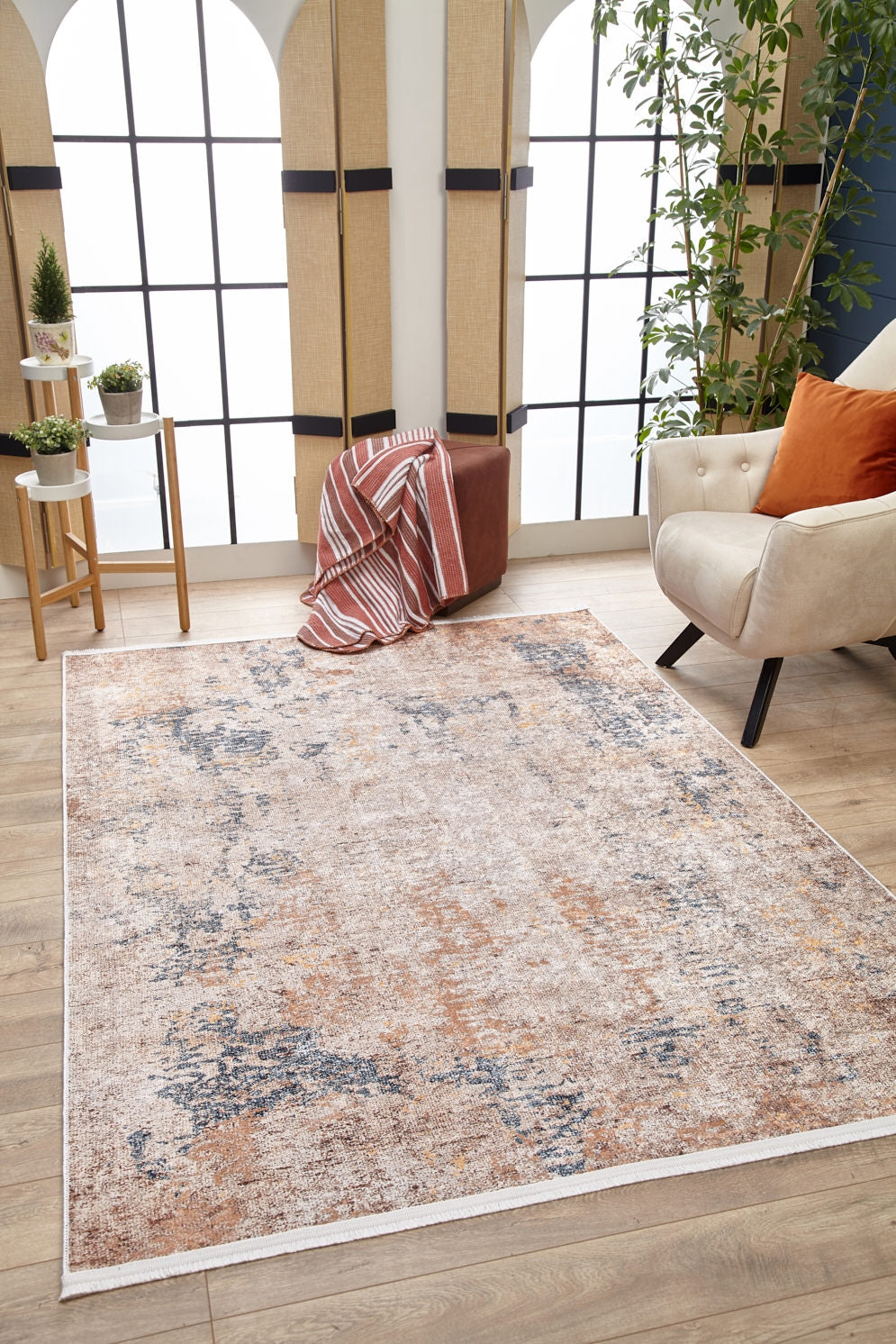 A modern living room featuring the Rhythmic Ripples - Washable Rug - JR1637 (Custom Size) in cream and beige, a beige armchair with an orange pillow, a brown ottoman with a striped blanket, a plant, and large windows bright with natural light.