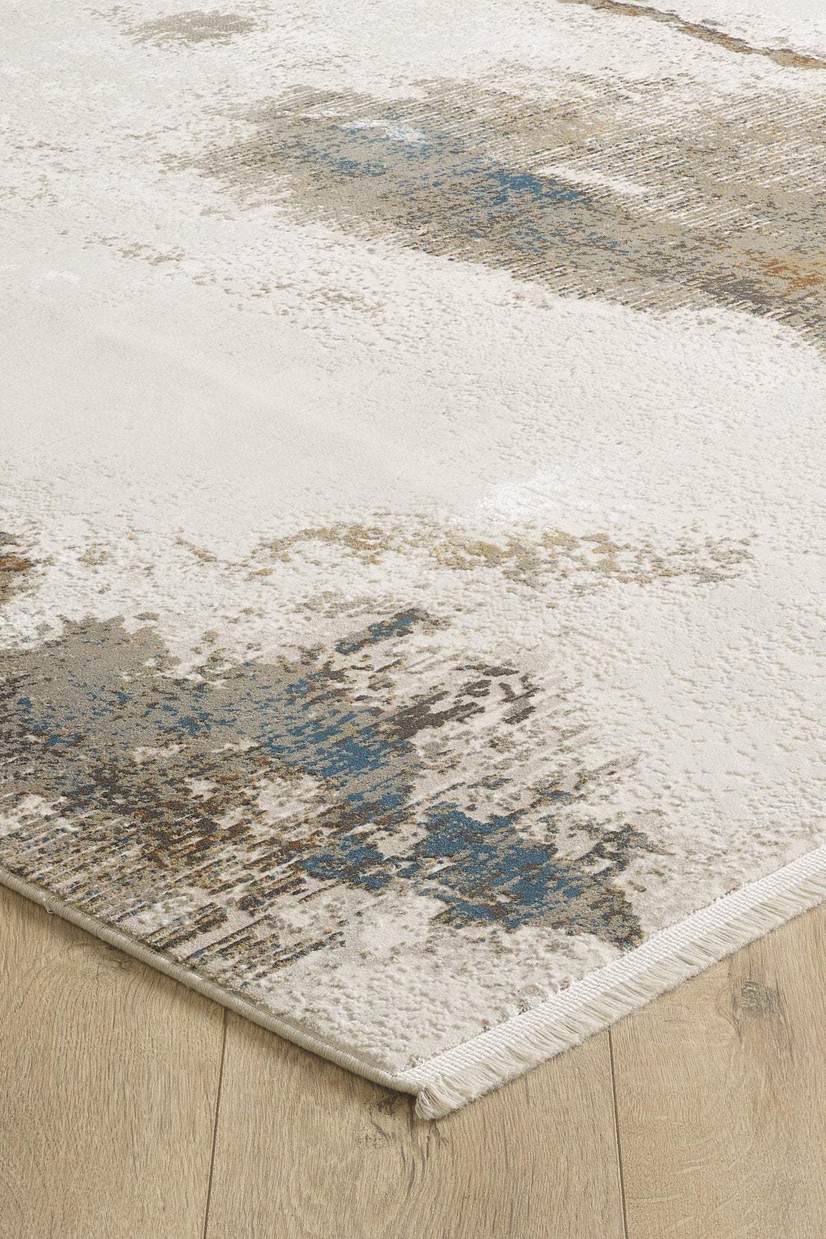 A close-up of the Riverstone Reflections Premium Area Rug - M685L, a modern machine-made acrylic-viscose rug with an abstract cream, blue, and brown design on a light wood floor. Features soft texture, finished edges, and short white fringe.
