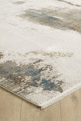 A close-up of the Riverstone Reflections Premium Area Rug - M685L, a modern machine-made acrylic-viscose rug with an abstract cream, blue, and brown design on a light wood floor. Features soft texture, finished edges, and short white fringe.