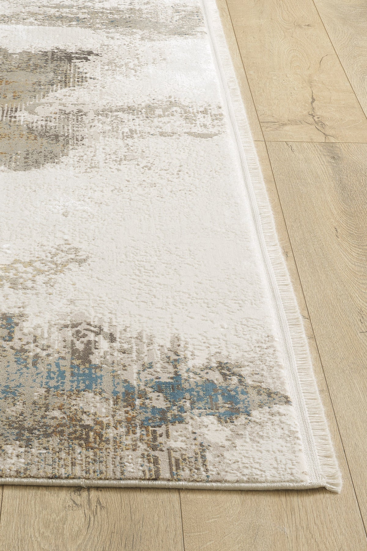A close-up of the Riverstone Reflections Premium Area Rug - M685L shows its soft texture, neat edges, and abstract beige, brown, and blue patterns. The machine-made rug is placed on a light wood floor.