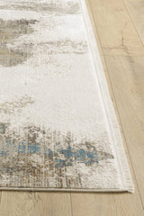 A close-up of the Riverstone Reflections Premium Area Rug - M685L shows its soft texture, neat edges, and abstract beige, brown, and blue patterns. The machine-made rug is placed on a light wood floor.