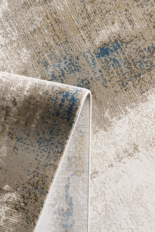 Load image 1 in gallery view. A close-up of the Riverstone Reflections Premium Area Rug - M685L, showcasing abstract patterns in beige, cream, blue, and gold with the corner folded over to display both front and back textures.