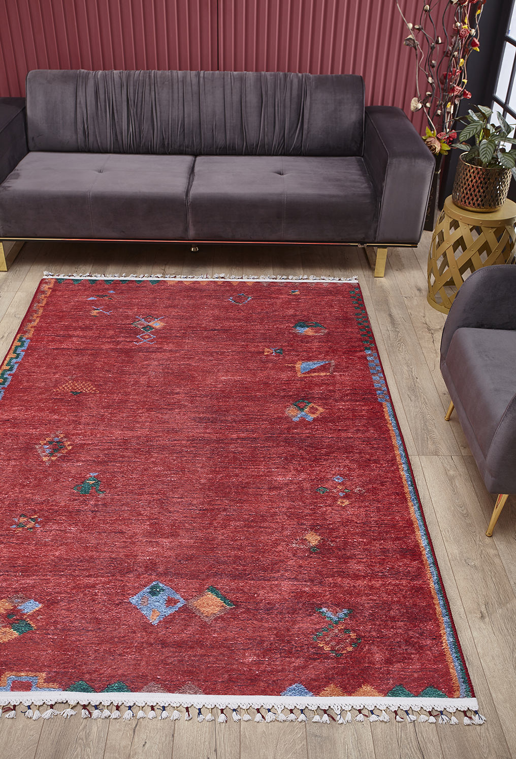 A living room features the Rosy Blush Machine-Washable Rug (JRS5067, custom size) in red with colorful geometric patterns. A dark gray sofa sits by vertical red panels, and a potted plant stands next to a wooden side table—ideal for high-traffic areas.