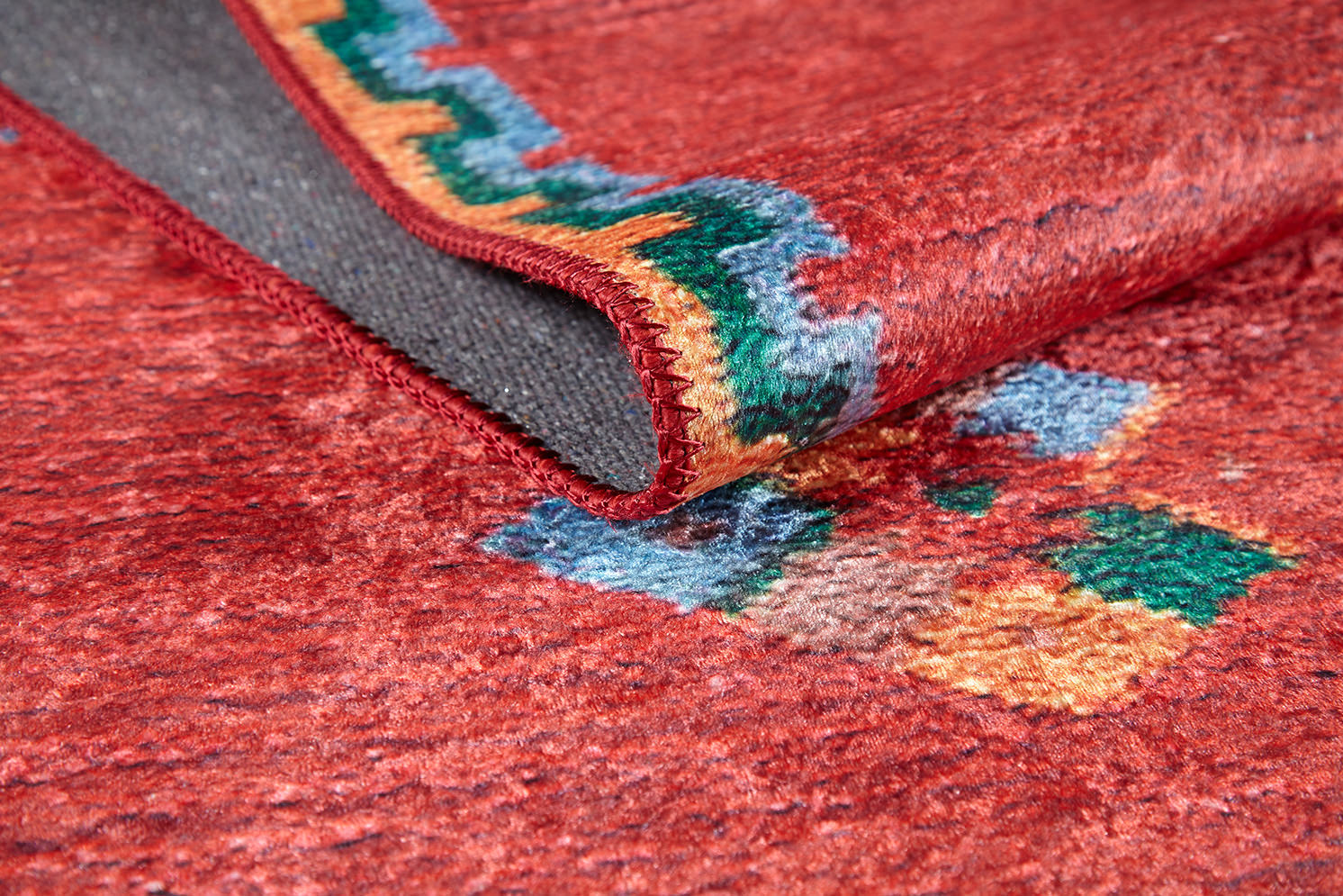 A close-up of the Rosy Blush - Machine-Washable Rug (JRS5067) in red, featuring colorful geometric patterns and a folded corner revealing its stitched edge and gray underside—perfect for high-traffic areas.