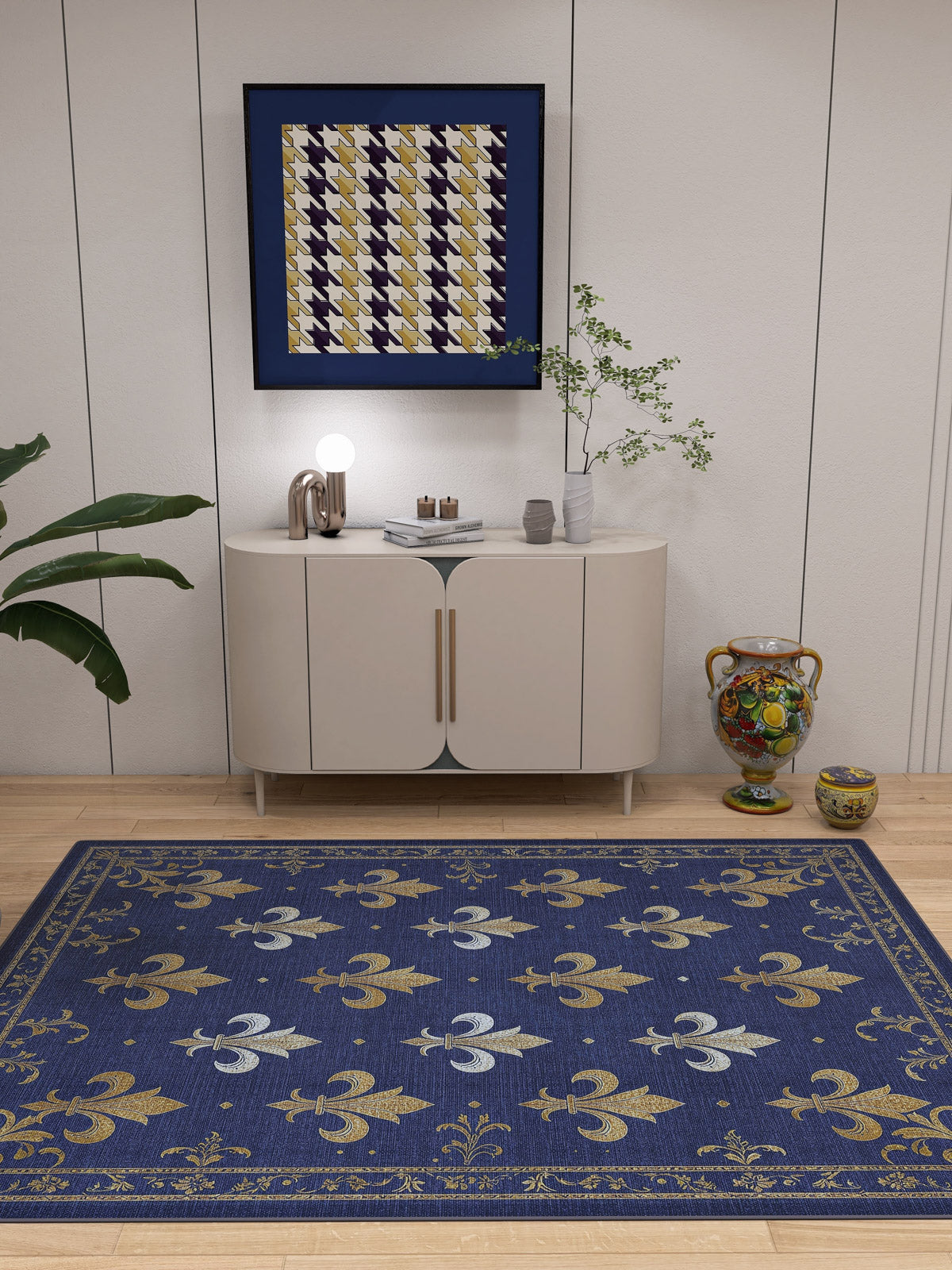 A modern room with a white cabinet, potted plants, and colorful vases features the Royal Fleur - Washable Rug - JRS016 (Custom Size) in blue, gold, and white fleur-de-lis—ideal for high-traffic or allergy-sensitive homes. Abstract art hangs above the cabinet.