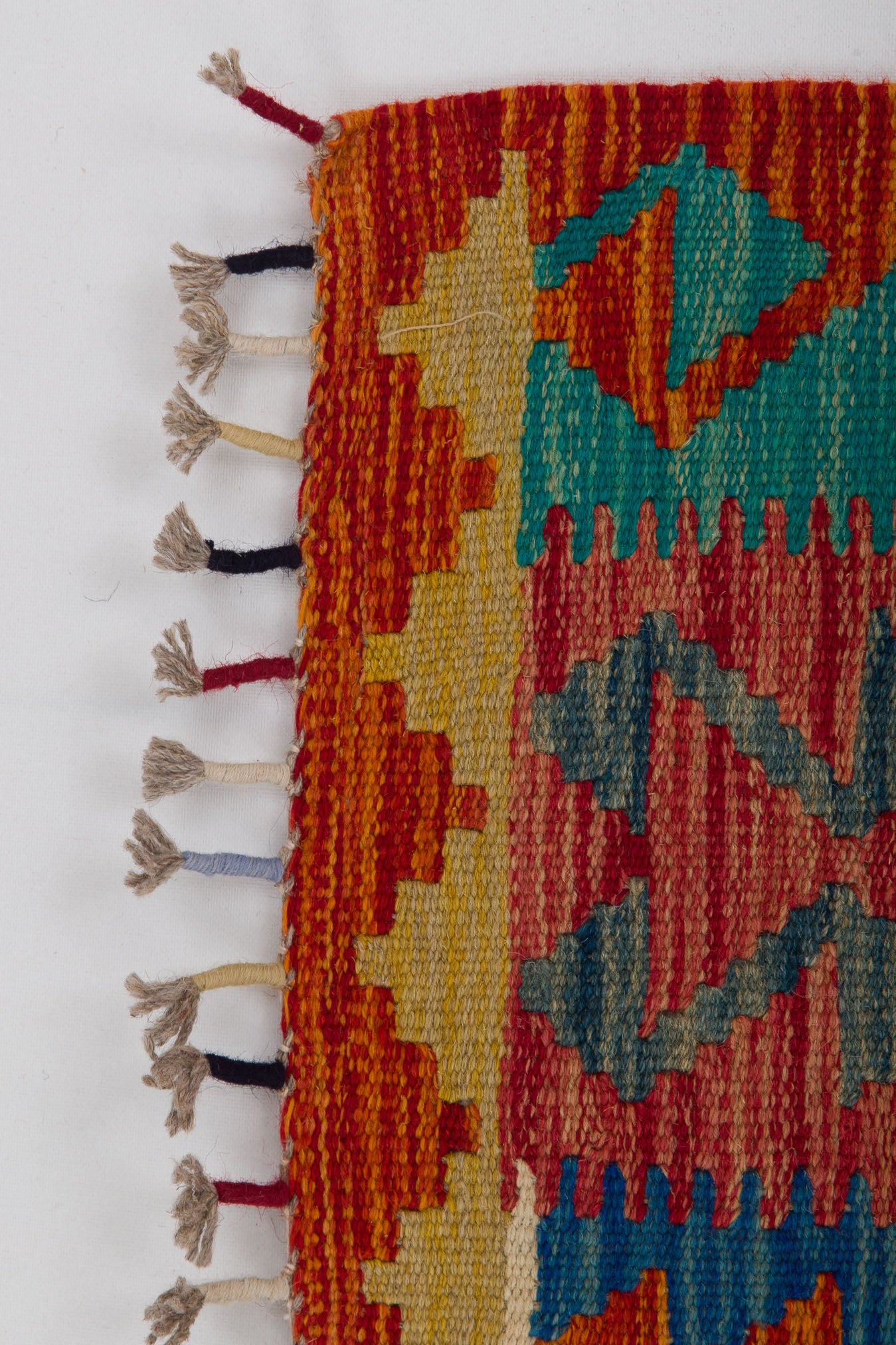 A close-up of the Royal Weft - Handwoven Kilim, showcasing colorful geometric patterns in red, orange, blue, and green wool with fringed tassels along the edge on a white background.
