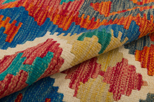 Load image 1 in gallery view. A close-up of the Royal Weft - Handwoven Kilim displays vibrant zigzag and geometric patterns in red, blue, yellow, green, and cream, with part of the flatweave rug folded to highlight its textured craftsmanship.