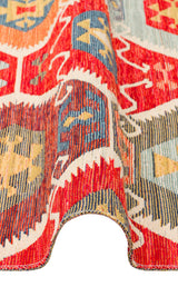 A close-up of the Ruby Grain - Kilim - NMD06 rug, featuring red, yellow, blue, and cream tribal geometric patterns, shown with a wavy folded edge to highlight its texture and thickness.