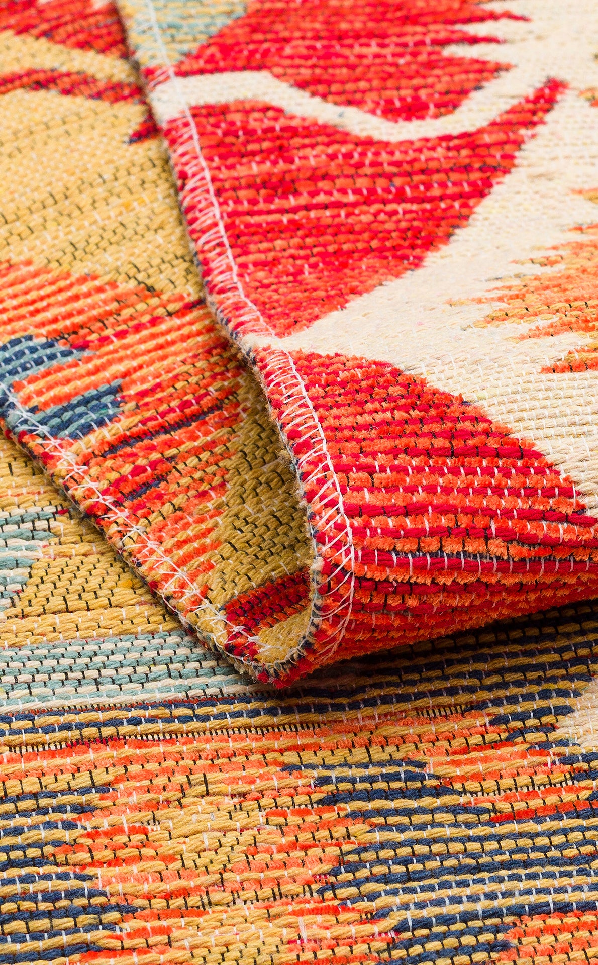 Close-up of the Ruby Grain - Kilim - NMD06 rug, showcasing colorful red, yellow, blue, and cream tribal patterns with visible stitching. A folded corner reveals the flatwoven underside and intricate texture.