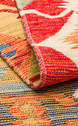 Close-up of the Ruby Grain - Kilim - NMD06 rug, showcasing colorful red, yellow, blue, and cream tribal patterns with visible stitching. A folded corner reveals the flatwoven underside and intricate texture.