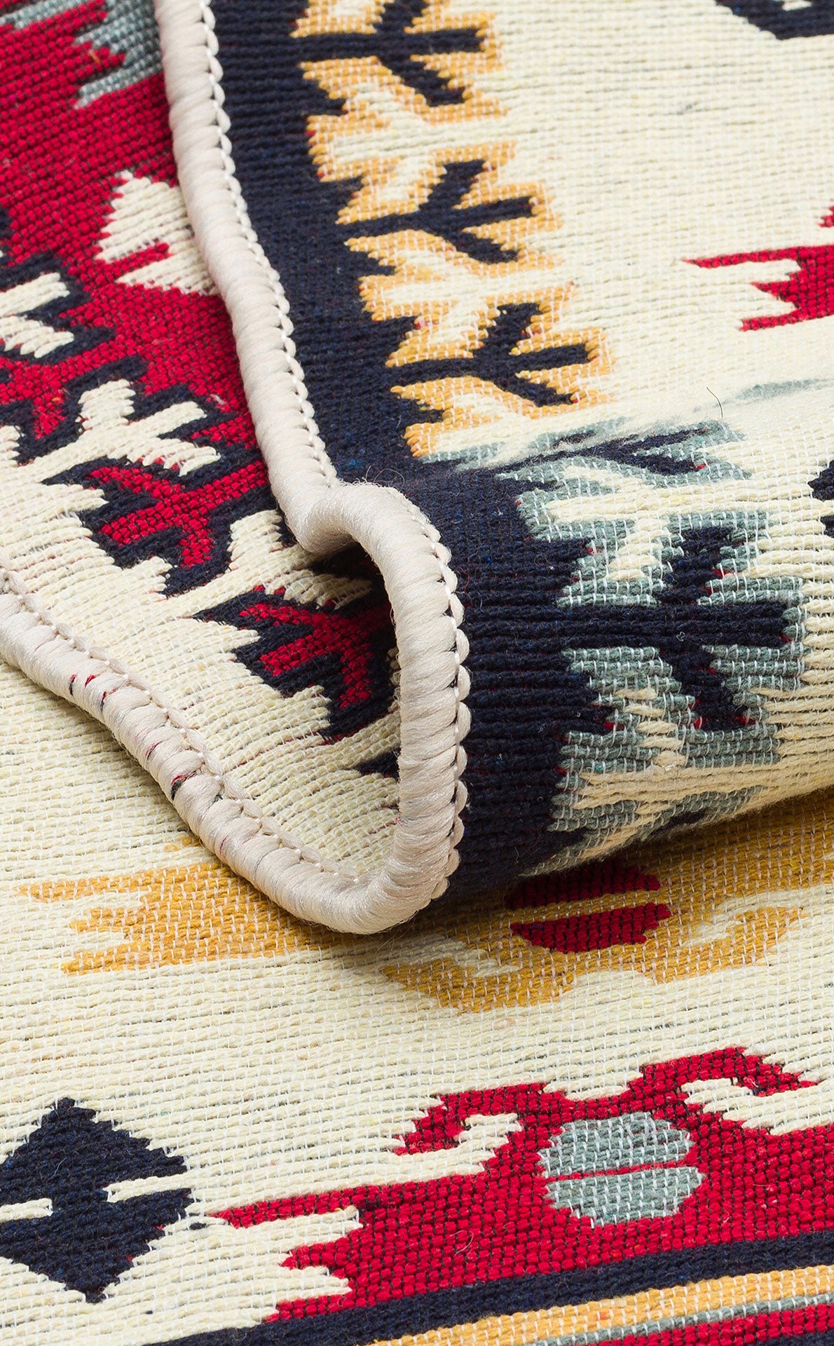 A close-up of Ruby Weft - Washable Double-Sided Kilims - AR30, partially folded to showcase its detailed stitching and bold geometric patterns in red, yellow, black, and ivory along the edge.