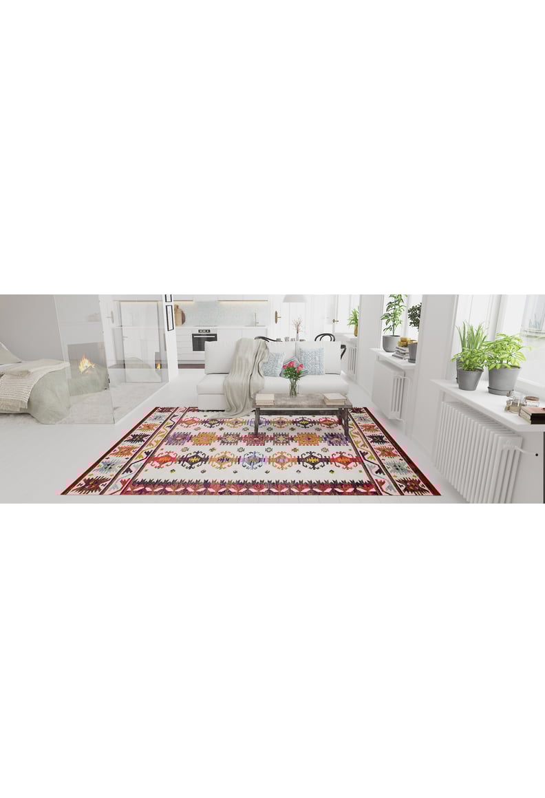 A bright living room with white walls and furniture features the Rustic Refrain - Washable Rug - JR149, green plants on the windowsills, a fireplace in the background, and a cushioned bench with a light throw blanket centered on the rug.