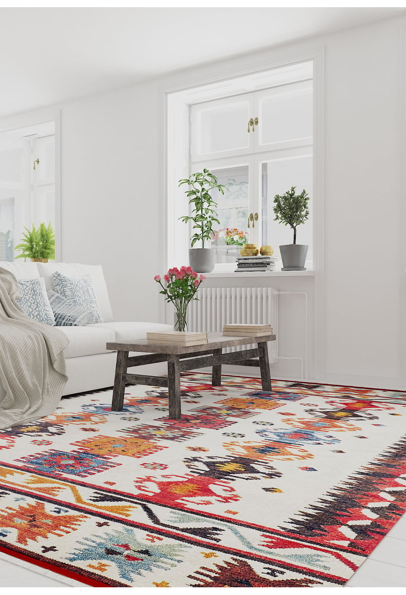 A bright, modern living room features the Rustic Refrain - Washable Rug - JR149 (Custom Size), a white sofa with blue pillows, a wooden coffee table with flowers, and potted plants on the windowsill in front of large windows.