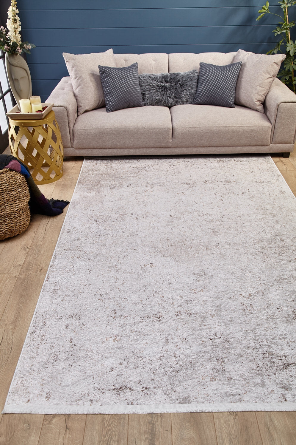A cozy living room with a beige sofa, gray and cream pillows, a fluffy cushion, and the Rustic Trails - Washable Rug - JR1736 (Custom Size) perfect for high-traffic areas, plus a yellow side table, candles, plants, blanket basket on wooden flooring.