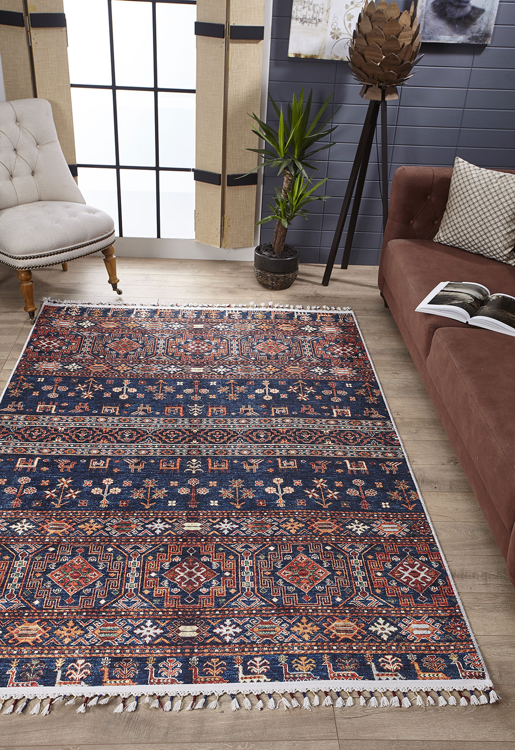 A living room features the Sahara Sunrise - Washable Rug - JR5091 (Custom Size) in blue, red, and cream on wood floors, paired with a brown sofa, beige armchair, potted plant, and large windows—perfect for allergy-sensitive rooms full of light.