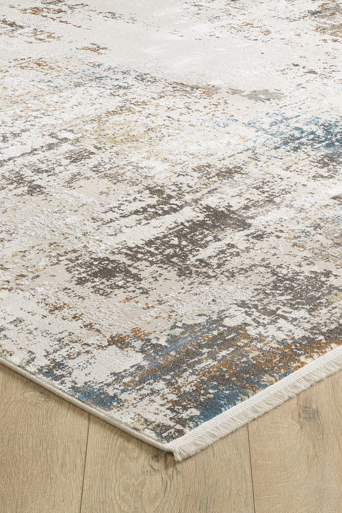 Load image 1 in gallery view. A close-up of the Sandy Papyrus Canvas Area Rug - M679L, a machine-made rug with a textured abstract pattern in beige, gray, blue, and gold tones, accented by a slight fringe and displayed on a light wood floor.