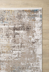 A partial view of the Sandy Papyrus Canvas Area Rug - M679L, featuring a machine-made abstract, marble-inspired pattern in brown, white, blue, and gold on a light wood floor with a fringed top edge.