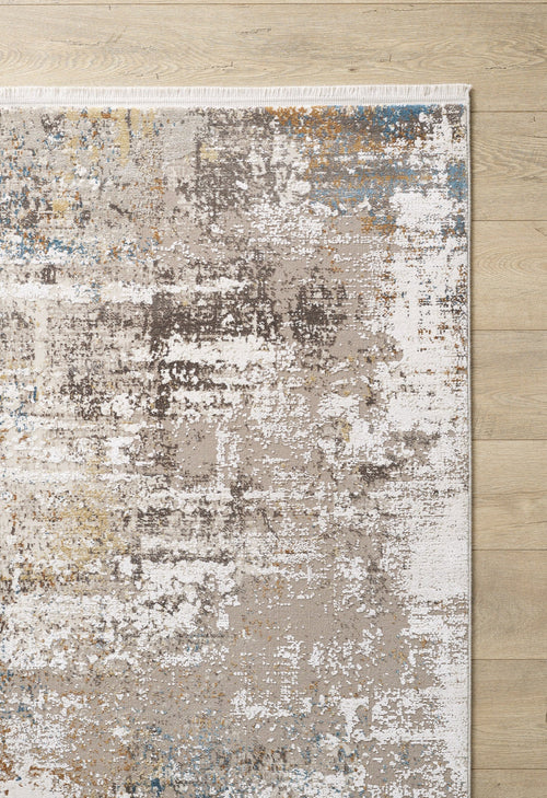 Load image 1 in gallery view. A partial view of the Sandy Papyrus Canvas Area Rug - M679L, featuring a machine-made abstract, marble-inspired pattern in brown, white, blue, and gold on a light wood floor with a fringed top edge.