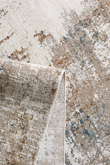 A close-up of the Sandy Papyrus Canvas Area Rug - M679L features abstract designs in beige, brown, white, and blue. A corner is folded over to showcase the rug's machine-made texture and thickness.