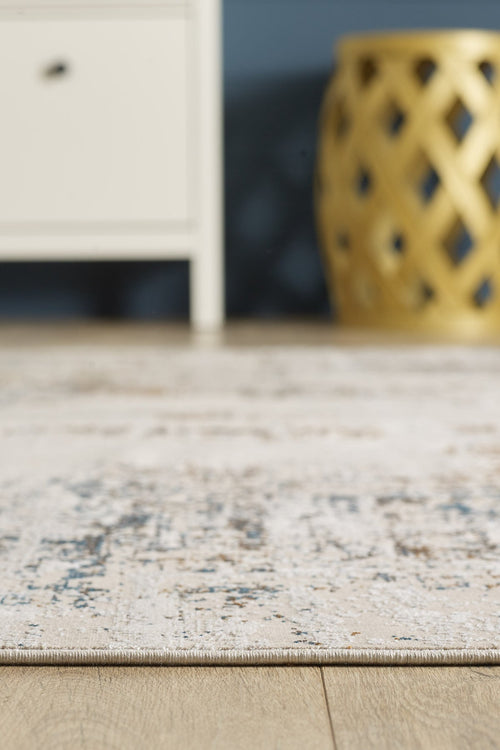 Load image 1 in gallery view. A close-up of the Sandy Papyrus Canvas Area Rug - M679L with subtle patterns on a wooden floor, placed beside a white cabinet and a gold lattice stool, set against a blue wall.