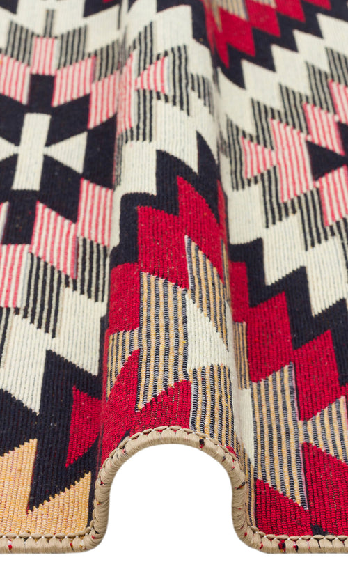 Load image 1 in gallery view. A close-up of the Scarlet Path - Washable Double-Sided Kilims - AR33, displaying its geometric red, black, cream, pink, and yellow pattern. The folded fabric reveals its thickness and detailed stitched edge.