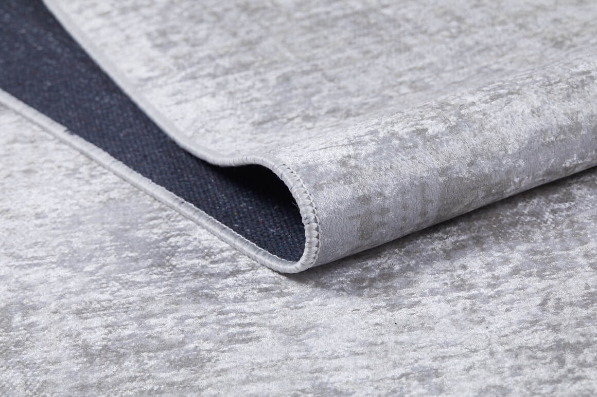 A close-up of the Sea Spray - Washable Rug - JR847 (Custom Size) in light gray shows its flat weave and a folded corner revealing a darker, non-slip backing.