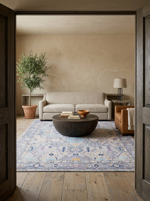 Load image 1 in gallery view. A cozy living room features a light beige sofa, tan armchair, and round wooden table. The Seraphix Pastel Bordered Washable Rug (LCC3011) covers the wood floor, while neutral walls and a potted tree enhance the inviting vibe.