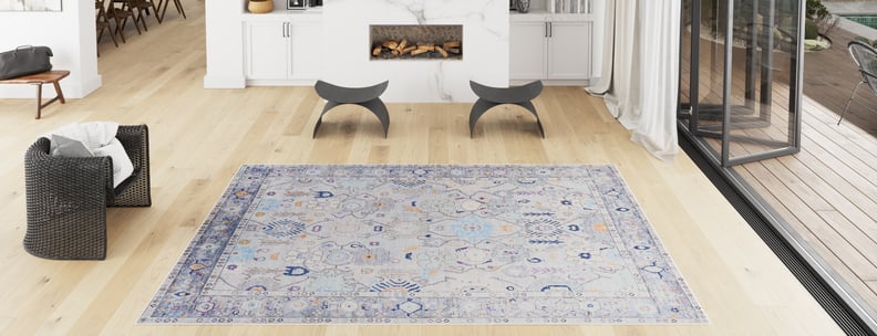 A modern living room with light wood floors, a Seraphix Pastel Bordered Washable Rug (LCC3011) in blue-gray hues, a black wicker chair, two curved stools, a fireplace, and large glass doors—perfect for high-traffic areas.