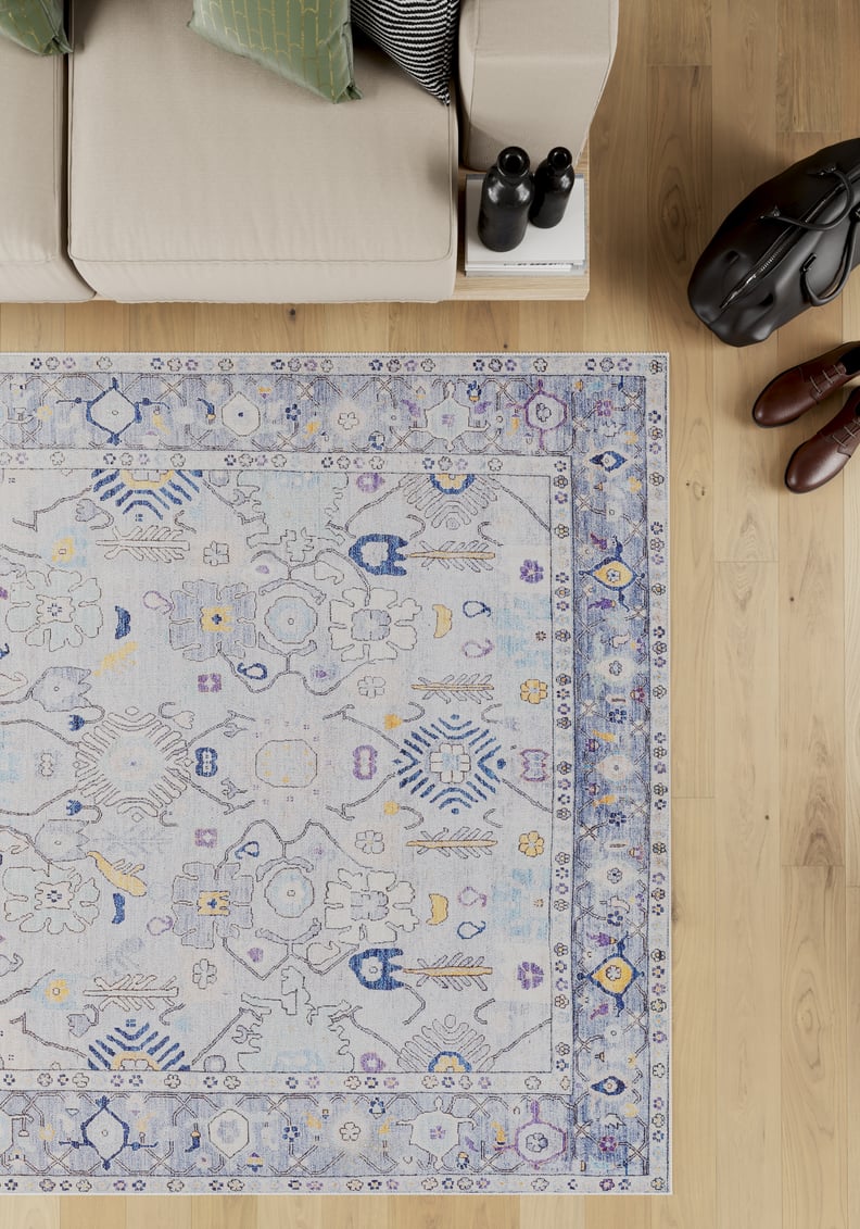 The Seraphix Pastel Bordered Washable Rug - LCC3011, with blue and yellow patterns, lies on a light wooden floor by a beige sofa—perfect for high-traffic areas. Nearby are books, binoculars, a black bag, and brown shoes.