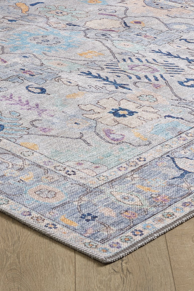 A close-up of the Seraphix Pastel Bordered Washable Rug - LCC3011 shows its blue base with muted geometric and floral patterns in lavender, yellow, and cream, lying on a wooden floor—perfect for high-traffic areas.